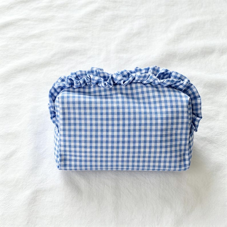 May include: A rectangular fabric pouch featuring a blue and white gingham check pattern. The top edge of the pouch is finished with a gathered ruffle trim made from the same blue and white checkered fabric, creating a soft, textured appearance.