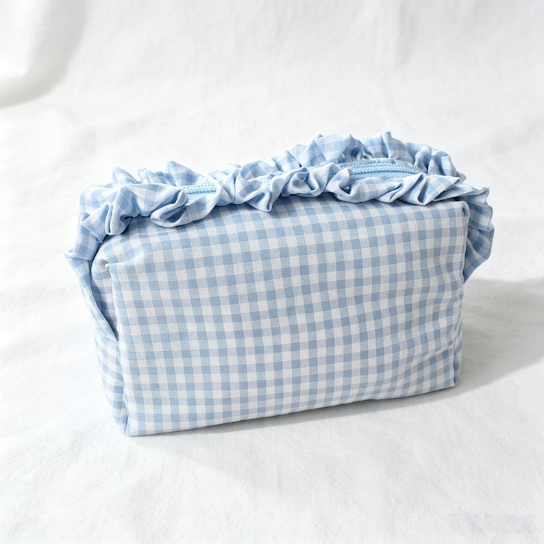 May include: A rectangular cosmetic pouch featuring a light blue and white gingham check pattern. The top of the pouch is accented with a gathered fabric ruffle trim that runs along the zip closure, which is also in a matching light blue colour.