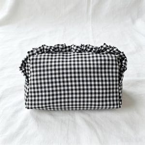 May include: A rectangular zipped pouch featuring a black and white gingham check pattern. The top edge of the pouch is finished with a gathered fabric ruffle that extends along the upper seam, providing a textured detail to the overall design.