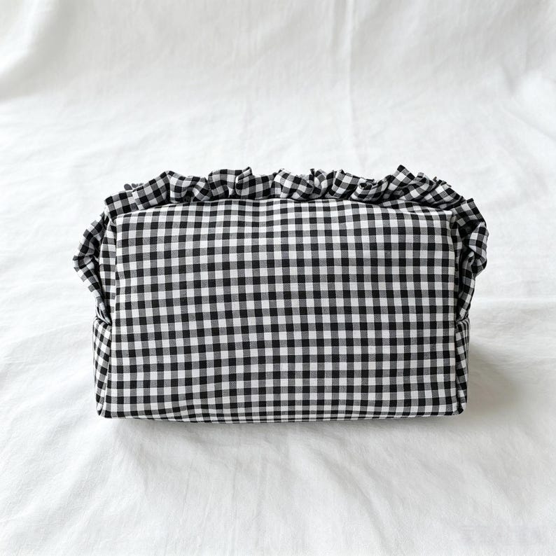 May include: A rectangular zipped pouch featuring a black and white gingham check pattern. The top edge of the pouch is finished with a gathered fabric ruffle that extends along the upper seam, providing a textured detail to the overall design.