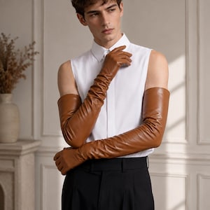 Peut inclure: Une paire de longs gants en cuir havane de style opéra qui dépassent les coudes. Les gants présentent une finition lisse et ajustée, portés avec une chemise blanche sans manches à col et un pantalon noir, offrant une allure structurée.
