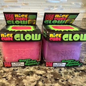 Puede incluir: Dos juguetes sensoriales Nee Doh Nice Cube Glow en su embalaje original. Una caja contiene un cubo rosa y la otra un cubo morado. Las cajas negras presentan texto verde brillante que dice Nice Cube Glow y Super Solid Squish, con una ventana.