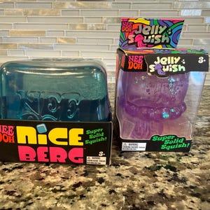 Könnte beinhalten: Zwei Nee Doh Sensorik-Spielzeuge in Originalverpackung. Links ein türkisfarbener, durchscheinender Nice Berg-Block in einer schwarzen Schachtel. Rechts eine violette, durchscheinende Jelly Squish-Quallenfigur in einer schwarzen Schachtel mit Karte.