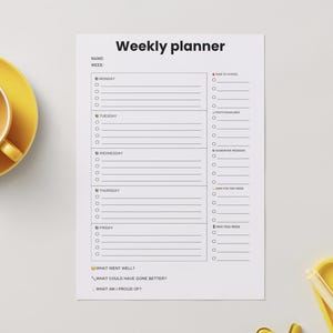 Op de afbeelding: Een witte weekplanner met zwarte tekst, een gele theekop en een geel dienblad. De planner heeft secties voor elke dag van de week, met selectievakjes en ruimte voor notities. De tekst "Weekly planner" staat bovenaan.