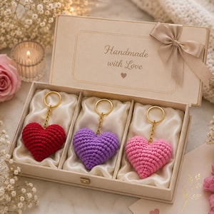May include: A beige gift box containing three crocheted heart keychains in red, purple, and pink. The box lid features the text "Handmade with Love" and a satin ribbon bow. Each keychain has a gold-toned metal ring and chain attached to the heart.
