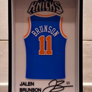 May include: A framed 3D display featuring a blue New York Knicks basketball jersey with the name BRUNSON and number 11 in orange and white. Below the jersey, the text JALEN BRUNSON is printed next to a black signature in a white shadow box frame.