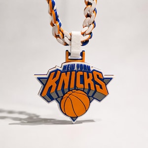 May include: A New York Knicks basketball team pendant featuring the orange, blue, and white team logo with a basketball graphic and the text NEW YORK KNICKS. The pendant hangs from a thick, chunky chain with alternating orange, blue, and white links.