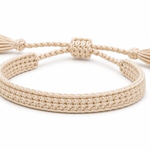 May include: A beige woven cord bracelet featuring a flat, textured band with a central sliding knot closure. The adjustable ends of the bracelet are finished with small, matching beige tassels, creating a cohesive and uniform appearance throughout.