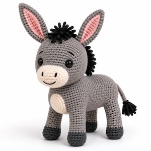 May include: A gray crocheted donkey toy with a cream-colored muzzle and belly, black hooves, and black mane. The donkey features large black safety eyes, a stitched smile, and tall ears with pink inner detailing. The tail ends in a black tassel.