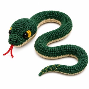 Pode incluir: Um brinquedo de cobra em crochê com a parte superior verde-escura e a parte inferior creme. A cobra tem olhos de plástico preto com detalhes bordados em amarelo, uma língua vermelha bifurcada e um pequeno padrão de ponto cruz amarelo perto da ponta da cauda.