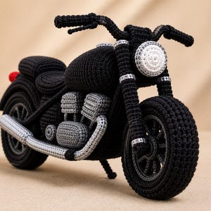 Może przedstawiać: Ręcznie wykonany szydełkowy motocykl z czarnym korpusem, białym okrągłym reflektorem i szarymi detalami silnika. Tekstylny model zawiera czarne siedzenie, kierownicę i koła, z małym czerwonym akcentem na tylnym błotniku, prezentując skomplikowane ściegi przędzy w całej strukturze.