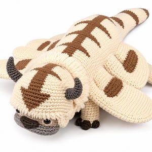 Op de afbeelding: Een crèmekleurige gehaakte knuffel in de vorm van een wezen met zes poten, bruine strepen op de rug en staart, een bruine pijl op de kop, donkergrijze gebogen hoorns, een grijze snuit en kleine donkere ogen, gemaakt met zichtbare steken.