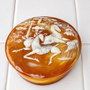 May include: A round, amber-colored lidded box featuring a carved white relief design on the top. The carving depicts two deer in a forest setting with trees and foliage. The surface has a smooth, glossy finish with translucent qualities.