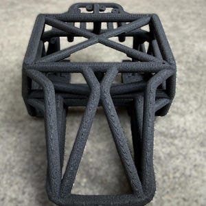 May include: A black 3D-printed chassis frame featuring a rigid, interconnected lattice structure with a rectangular top section and a tapered, angular base. The surface exhibits a distinct, layered texture characteristic of additive manufacturing.