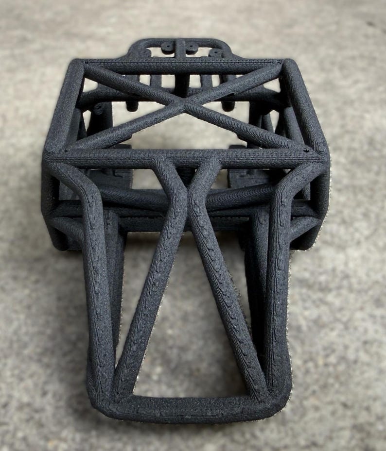 May include: A black 3D-printed chassis frame featuring a rigid, interconnected lattice structure with a rectangular top section and a tapered, angular base. The surface exhibits a distinct, layered texture characteristic of additive manufacturing.