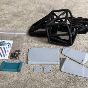 May include: A black 3D-printed roll cage frame for a remote-controlled vehicle, accompanied by several grey textured body panels, a small teal bracket, a clear plastic bag containing metal screws, and a sticker sheet featuring a flag design.