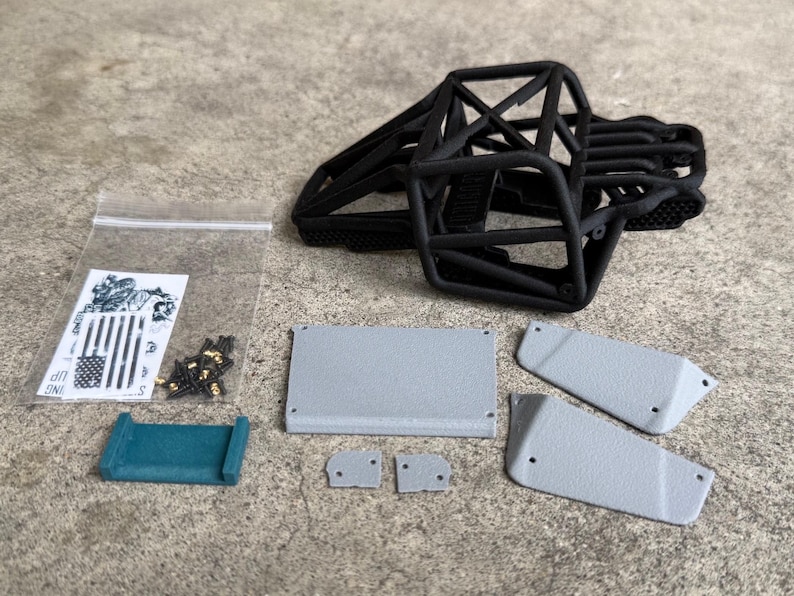 May include: A black 3D-printed roll cage frame for a remote-controlled vehicle, accompanied by several grey textured body panels, a small teal bracket, a clear plastic bag containing metal screws, and a sticker sheet featuring a flag design.