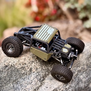 Puede incluir: Un vehículo de juguete a control remoto tipo rock crawler en color verde oliva con una jaula antivuelco negra, neumáticos negros con tacos y un panel de techo decorado con un gráfico de la bandera estadounidense en color blanco.