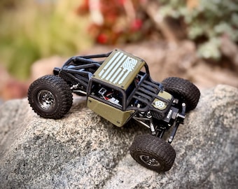 May include: A miniature olive green remote-controlled rock crawler vehicle featuring a black roll cage, knobby black tires, and a roof panel decorated with a white American flag graphic. The chassis is exposed with visible suspension components.