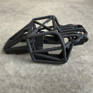 May include: A matte black 3D printed roll cage chassis for a remote control vehicle. The structure features a complex tubular frame design with a honeycomb pattern on the lower side panels and a central support beam across the top section.