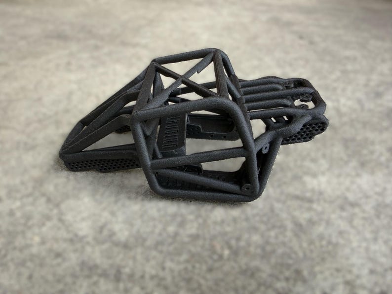 May include: A matte black 3D printed roll cage chassis for a remote control vehicle. The structure features a complex tubular frame design with a honeycomb pattern on the lower side panels and a central support beam across the top section.