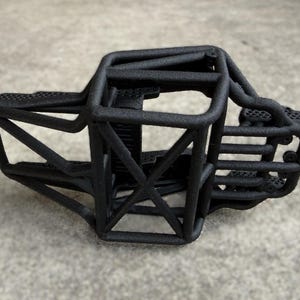 May include: A matte black 3D printed roll cage chassis for a remote control vehicle. The structure features a complex network of interconnected tubular bars forming a protective frame with a textured surface finish and open interior compartments.