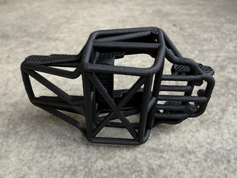 May include: A matte black 3D printed roll cage chassis for a remote control vehicle. The structure features a complex network of interconnected tubular bars forming a protective frame with a textured surface finish and open interior compartments.