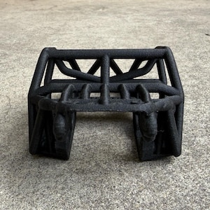 May include: A matte black, three-dimensional printed roll cage structure featuring a tubular frame design with a central horizontal bar, vertical supports, and an open-ended base, characterized by a textured surface finish throughout the entire object.