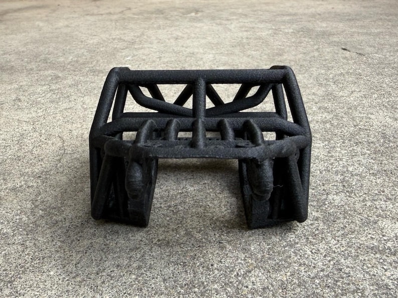 May include: A matte black, three-dimensional printed roll cage structure featuring a tubular frame design with a central horizontal bar, vertical supports, and an open-ended base, characterized by a textured surface finish throughout the entire object.