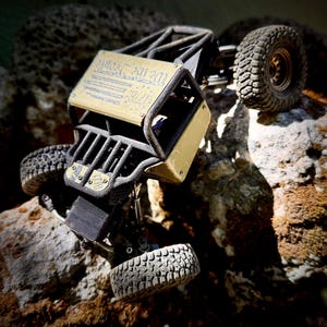 May include: A remote-controlled rock crawler vehicle with a black tubular frame and gold-colored body panels. The top panel features the text "We the People" in blue. The vehicle has four large, knobby black rubber tires and a detailed suspension system.
