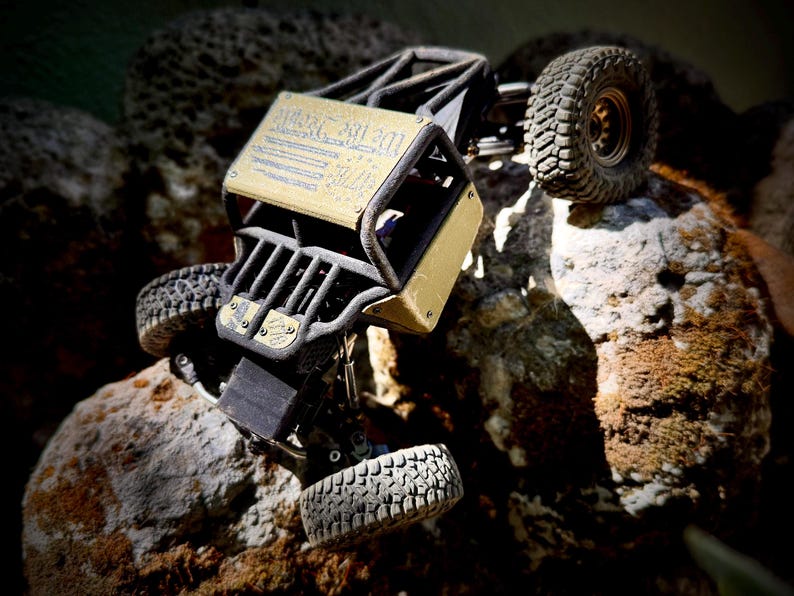 May include: A remote-controlled rock crawler vehicle with a black tubular frame and gold-colored body panels. The top panel features the text "We the People" in blue. The vehicle has four large, knobby black rubber tires and a detailed suspension system.