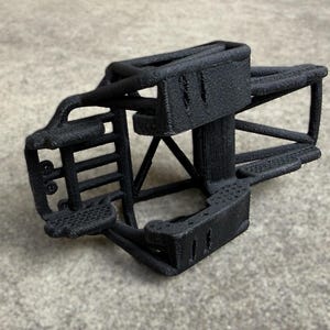 May include: A black 3D printed chassis frame for a remote control vehicle. The structure features a rigid, textured cage design with multiple support bars, a front bumper section, and mounting points for mechanical components and suspension parts.