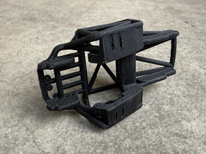 May include: A black 3D printed chassis frame for a remote control vehicle. The structure features a rigid, textured cage design with multiple support bars, a front bumper section, and mounting points for mechanical components and suspension parts.