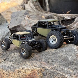 May include: Two olive green remote-controlled rock crawler vehicles with black roll cages and silver wheels. The vehicles feature exposed suspension components, large treaded tires, and a compact chassis design, positioned on a textured rock surface.