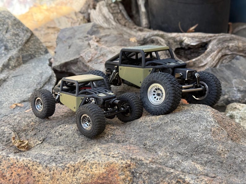 May include: Two olive green remote-controlled rock crawler vehicles with black roll cages and silver wheels. The vehicles feature exposed suspension components, large treaded tires, and a compact chassis design, positioned on a textured rock surface.