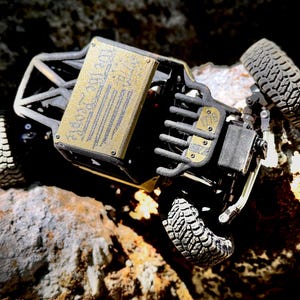 May include: A black remote-controlled rock crawler vehicle featuring a gold-colored metal roof panel engraved with the text "We The People" and a smaller gold-colored front plate engraved with "1776". The vehicle has four knobby rubber tires.