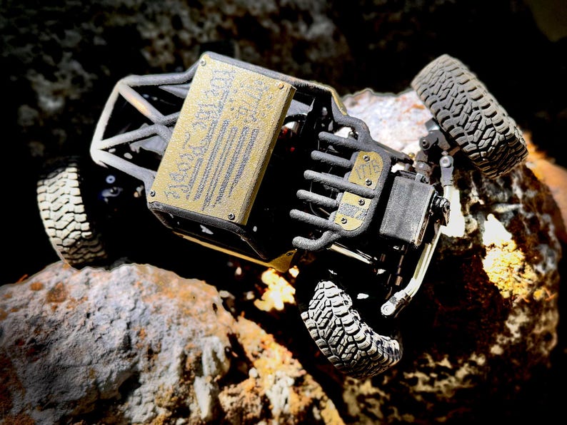 May include: A black remote-controlled rock crawler vehicle featuring a gold-colored metal roof panel engraved with the text "We The People" and a smaller gold-colored front plate engraved with "1776". The vehicle has four knobby rubber tires.