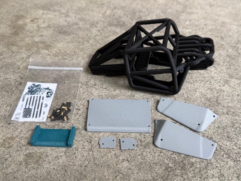 May include: A black 3D-printed roll cage frame for a remote-controlled vehicle, accompanied by several grey textured body panels, a small teal bracket, a clear plastic bag containing metal screws, and a sticker sheet featuring a flag design.