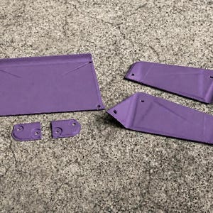 May include: A set of five purple, flat, rigid components with pre-drilled holes. The collection includes one large rectangular piece, two elongated trapezoidal panels, and two small, rounded square connectors, all featuring a matte purple finish.