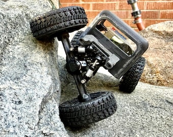 May include: A black remote-controlled crawler vehicle with large, deep-tread rubber tires and visible suspension shocks. The vehicle features a dark frame and a window decal with the text BFGoodrich, positioned at an angle against a large rock.