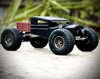 May include: A custom toy truck featuring a matte black cab with a low-profile roof, a dark red slatted cargo bed, and oversized black off-road tires with bronze-colored multi-spoke rims. The vehicle has a visible suspension and chassis.