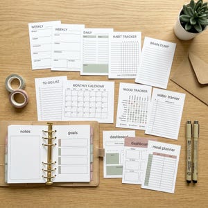 May include: A selection of planner inserts with headings such as 'Weekly', 'Daily', and 'Habit Tracker'. The inserts are white with minimalist text and pastel accents. A tan binder, pens, and decorative tape are also present. The image is set on a wooden surface.