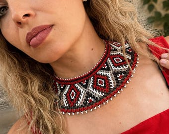 Peut inclure: Un collier ras-du-cou orné d'un motif géométrique complexe composé de petites perles rouges, noires, blanches et grises. Le design présente des losanges répétitifs et des lignes travaillées, avec une fine frange de perles sur le bord inférieur.