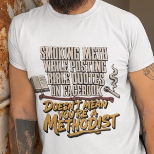 May include: A white short-sleeved t-shirt featuring a graphic print with the text "SMOKING METH WHILE POSTING BIBLE QUOTES ON FACEBOOK DOESN'T MEAN YOU'RE A METHODIST". The design includes an illustration of an open Bible and a smoking pipe.