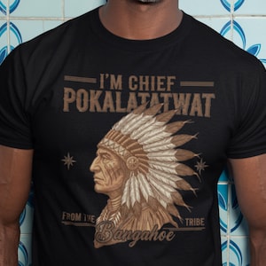 May include: A black short-sleeved t-shirt featuring a graphic print of a person wearing a feathered headdress in profile. The text reads I'M CHIEF POKALATATWAT FROM THE TRIBE Bangahoe in a tan, distressed font with two small star icons on the sides.