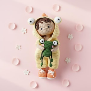 May include: A crocheted doll with brown hair wearing a yellow hooded frog-themed onesie and orange trainers. The doll holds a small green crocheted frog toy. The doll has black eyes and a simple stitched mouth, crafted with detailed yarn textures.