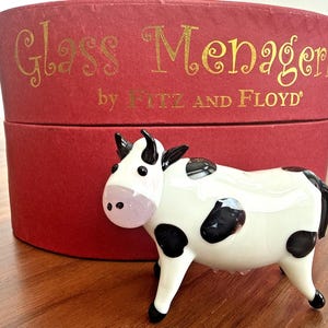May include: A white and black glass cow figurine with a pink nose and black spots. The cow is in front of a red box with gold text that reads "Glass Menagerie by FITZ AND FLOYD". The figurine is on a wooden surface.