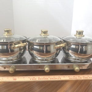 May include: A triple buffet server featuring three stainless steel pots with glass lids and gold-tone handles, set on a rectangular warming base with three gold-tone control knobs, positioned behind a wooden ruler measuring 24 inches in length.