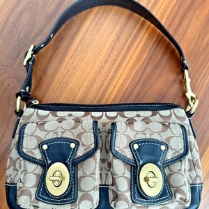May include: A brown and tan handbag with a repeating pattern. The bag has two front pockets with navy blue leather accents and gold-tone hardware. A dark brown leather strap arches over the top.