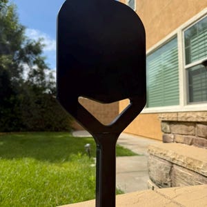 Könnte beinhalten: Eine schwarze Pizzaschieber mit rechteckiger Klinge und einem Griff mit dreieckigem Ausschnitt. Der Metallschieber steht auf einer Steinoberfläche, mit einem grünen Rasen und einem Haus im Hintergrund.